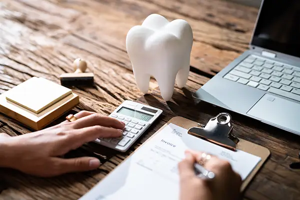 A man sitting at a desk with a calculator, laptop, and giant plastic tooth filling out insurance paperwork. A man sitting at a desk with a calculator, laptop, and giant plastic tooth filling out insurance paperwork.